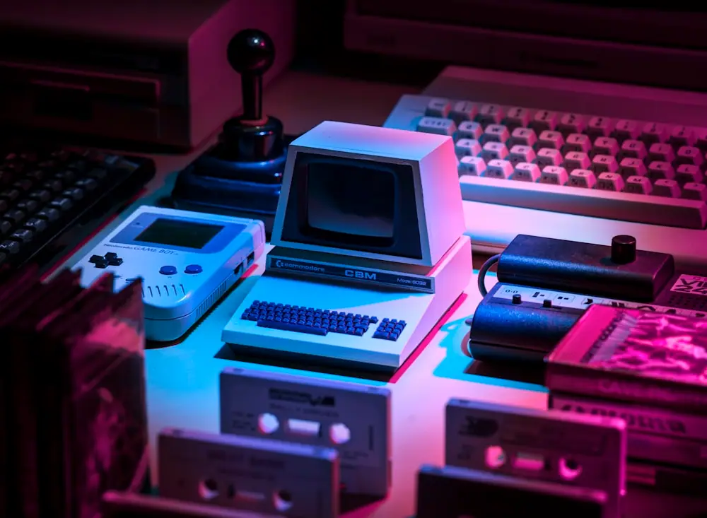 Consolas portáteis e computadores retro numa mesa iluminada a néon